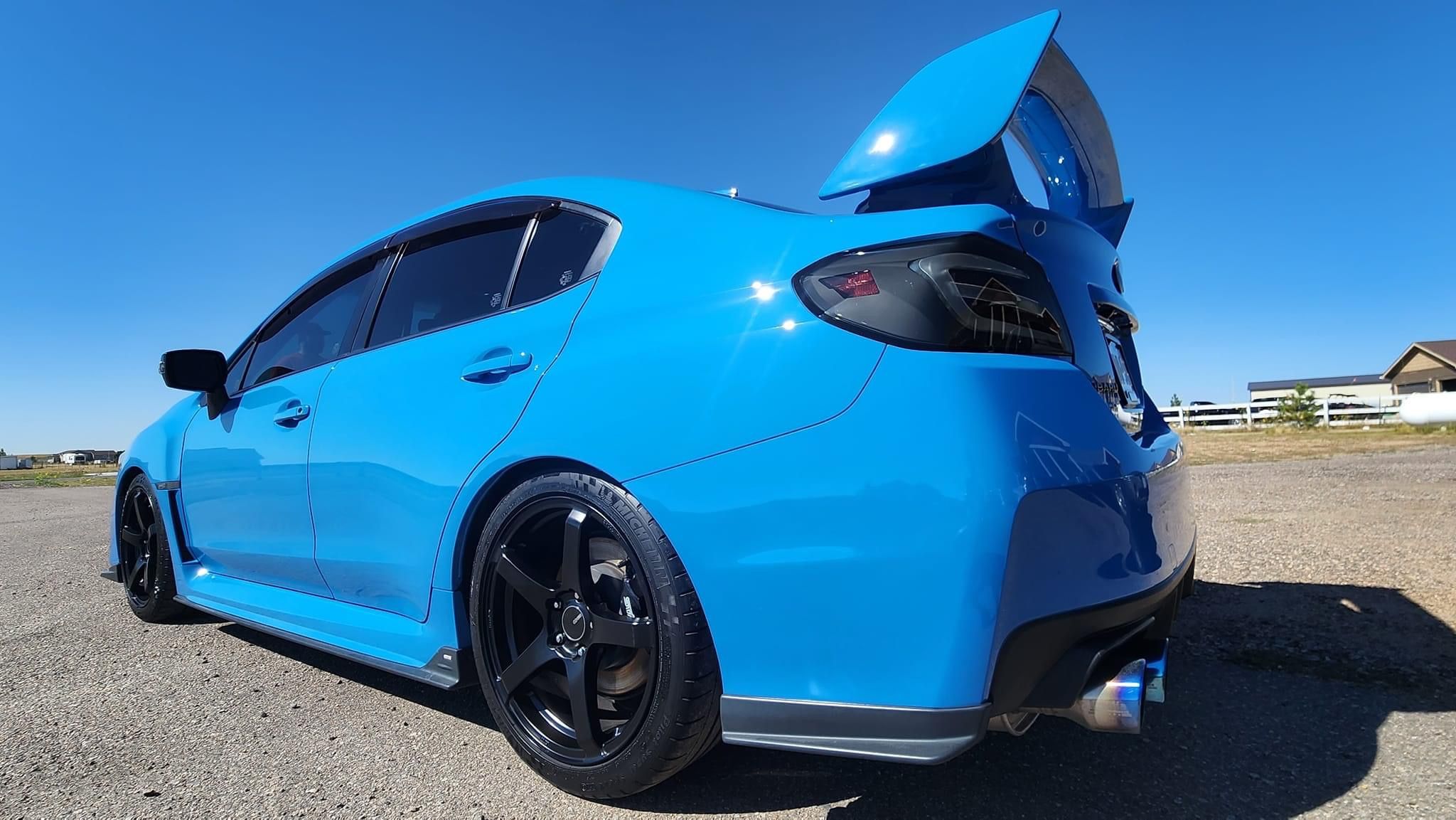 Blue Subaru sports car with black rims and large spoiler parked outdoors on a sunny day.
