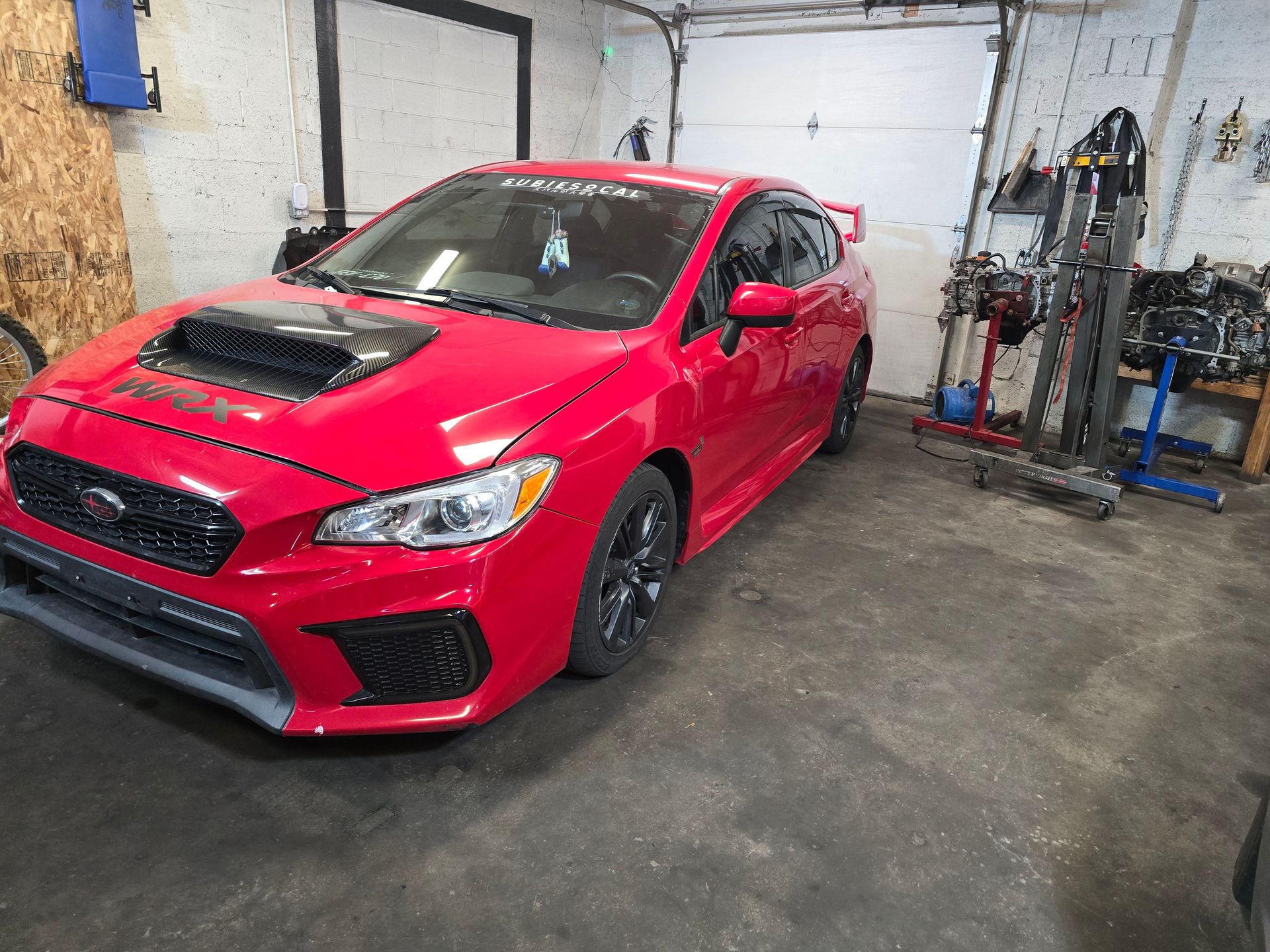 Red Subaru WRX in a garage. Black hood scoop, spoiler, and wheels.