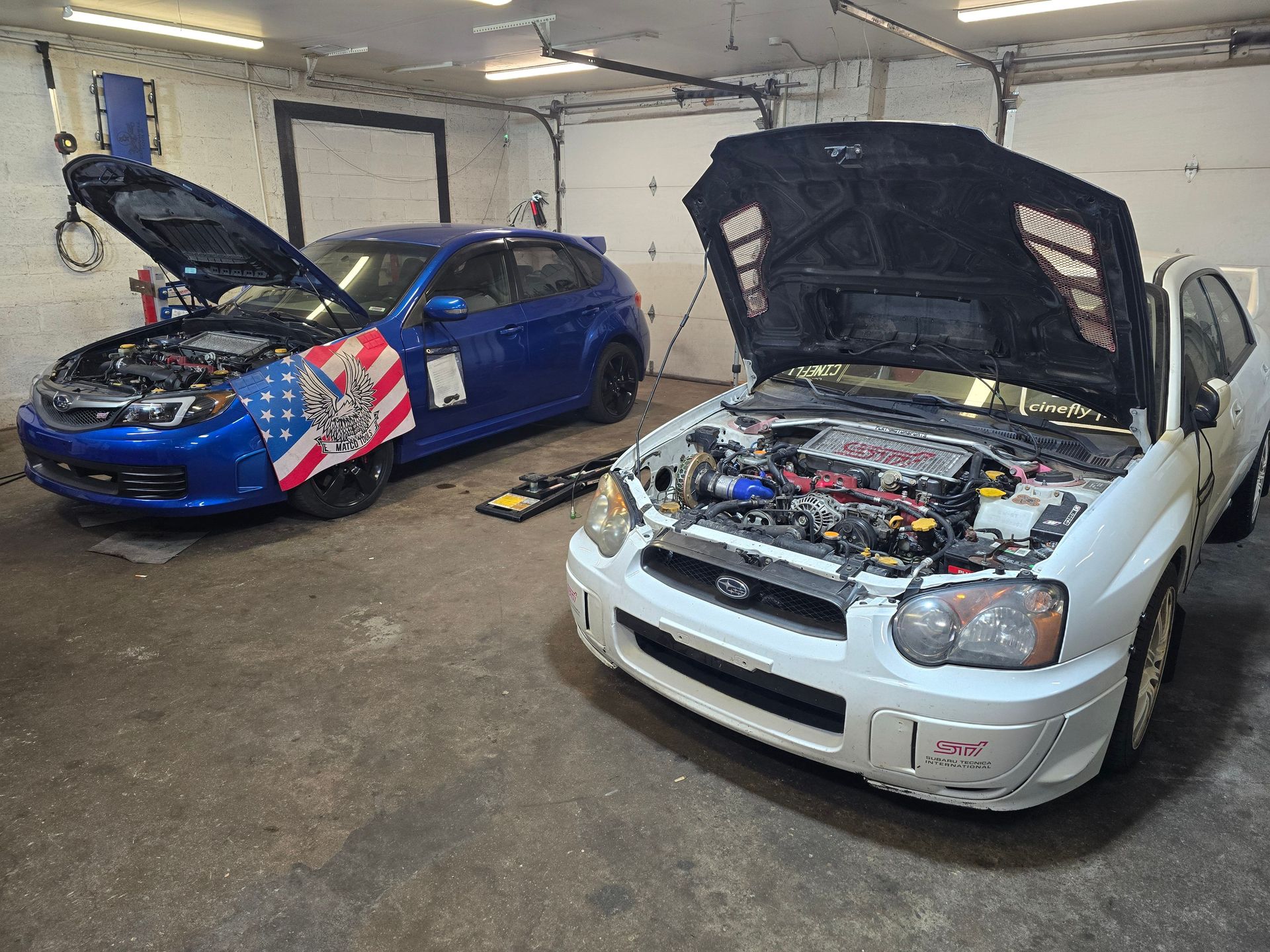Two Subaru WRX cars with open hoods inside a garage; one blue, one white.