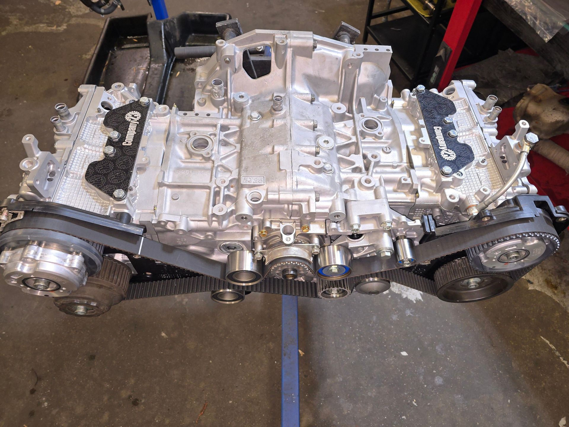 Silver V6 engine block with black timing belts in a garage setting.
