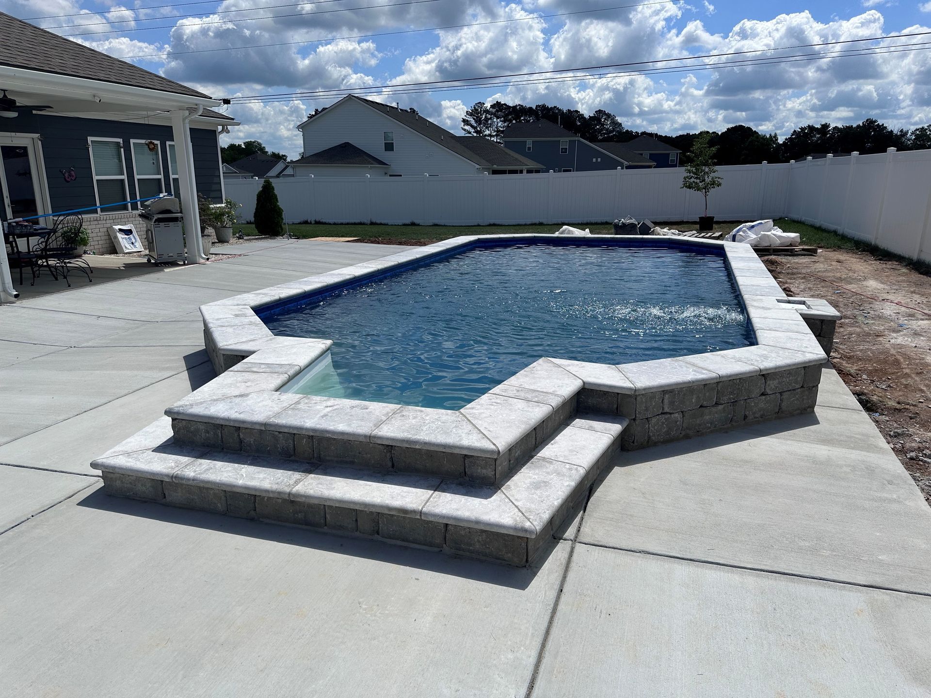 Backyard swimming pool with raised stone surround and concrete patio under a partly cloudy sky