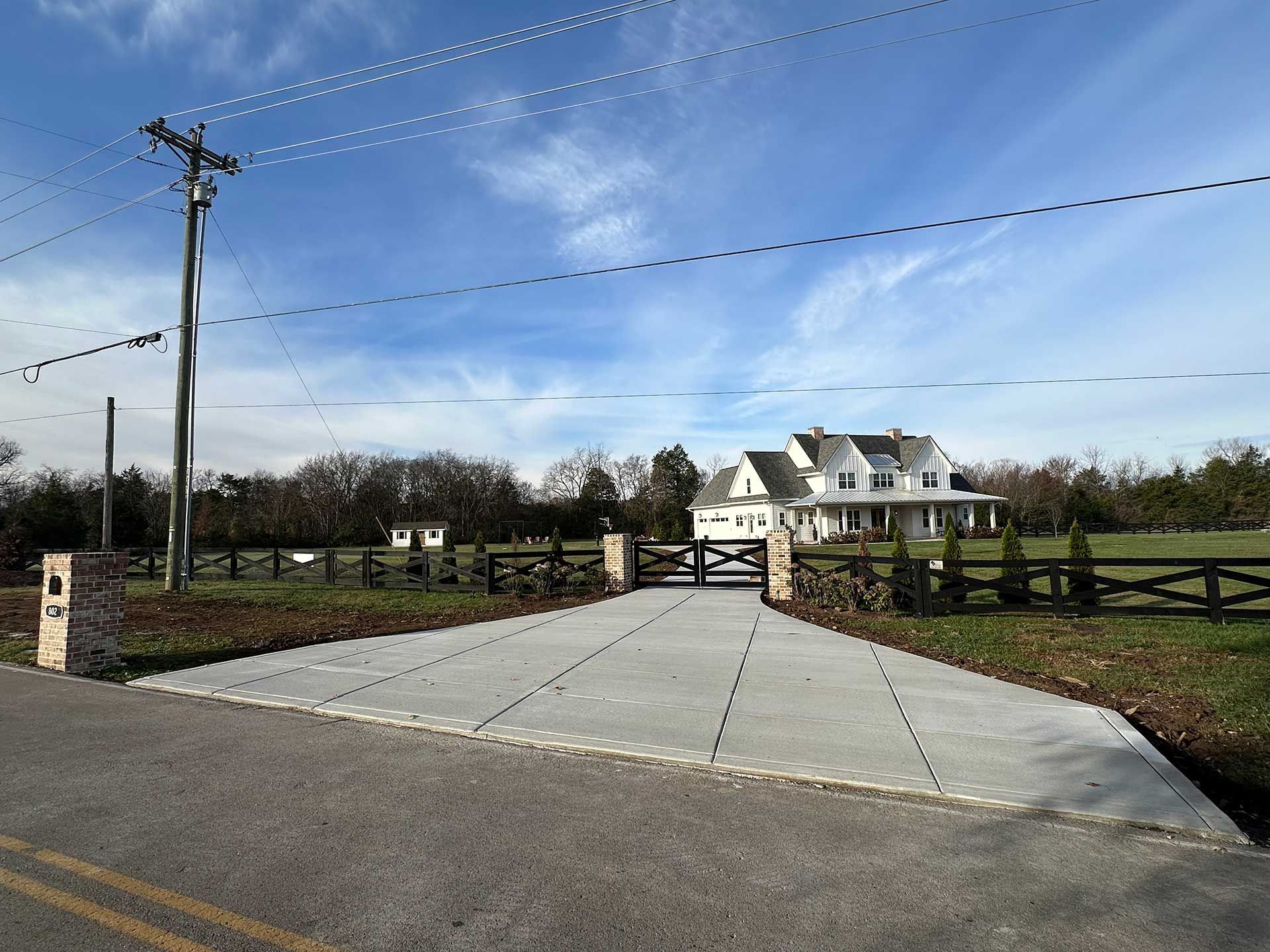 concrete driveway leading to a white farmhouse with black fencing under a partly cloudy blue sky