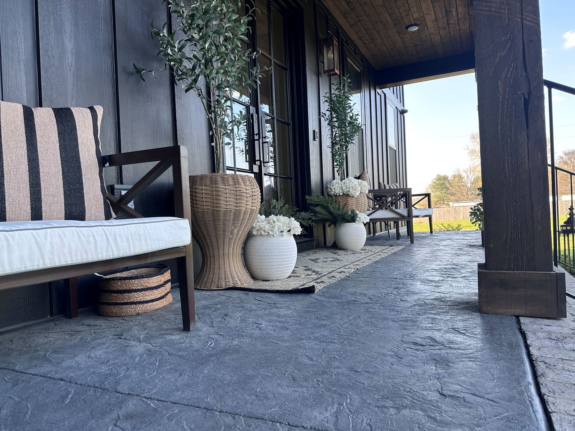 Porch with black-framed bench, striped cushion, woven baskets, potted plants, and stone floor in sunlight
