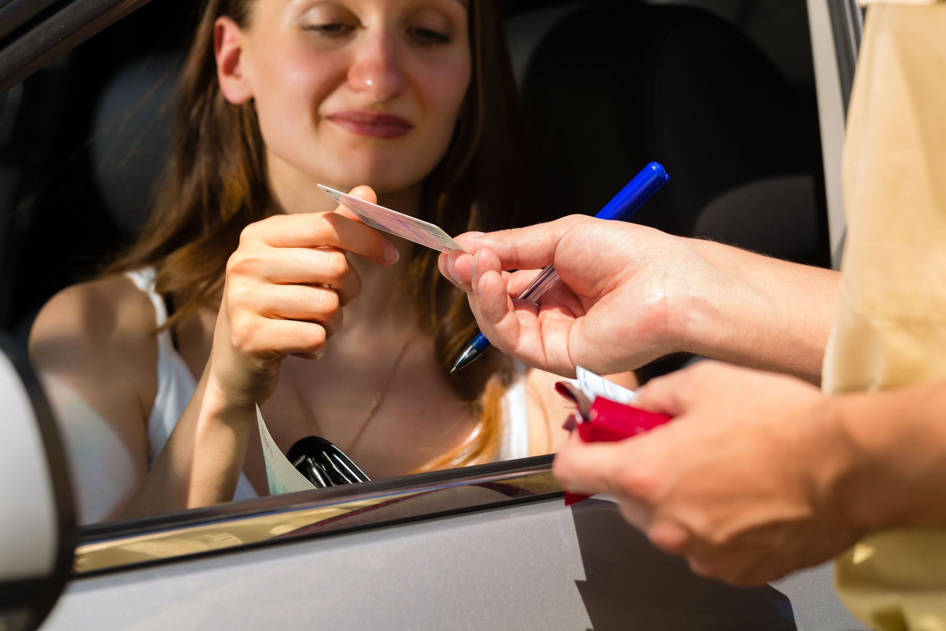 Woman in car receiving a document and pen from a person. Sunlight.