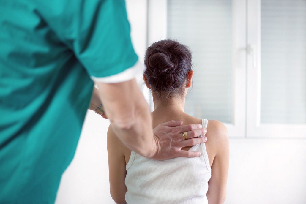Caregiver in teal shirt rests a hand on a seated person’s shoulder near a bright window.