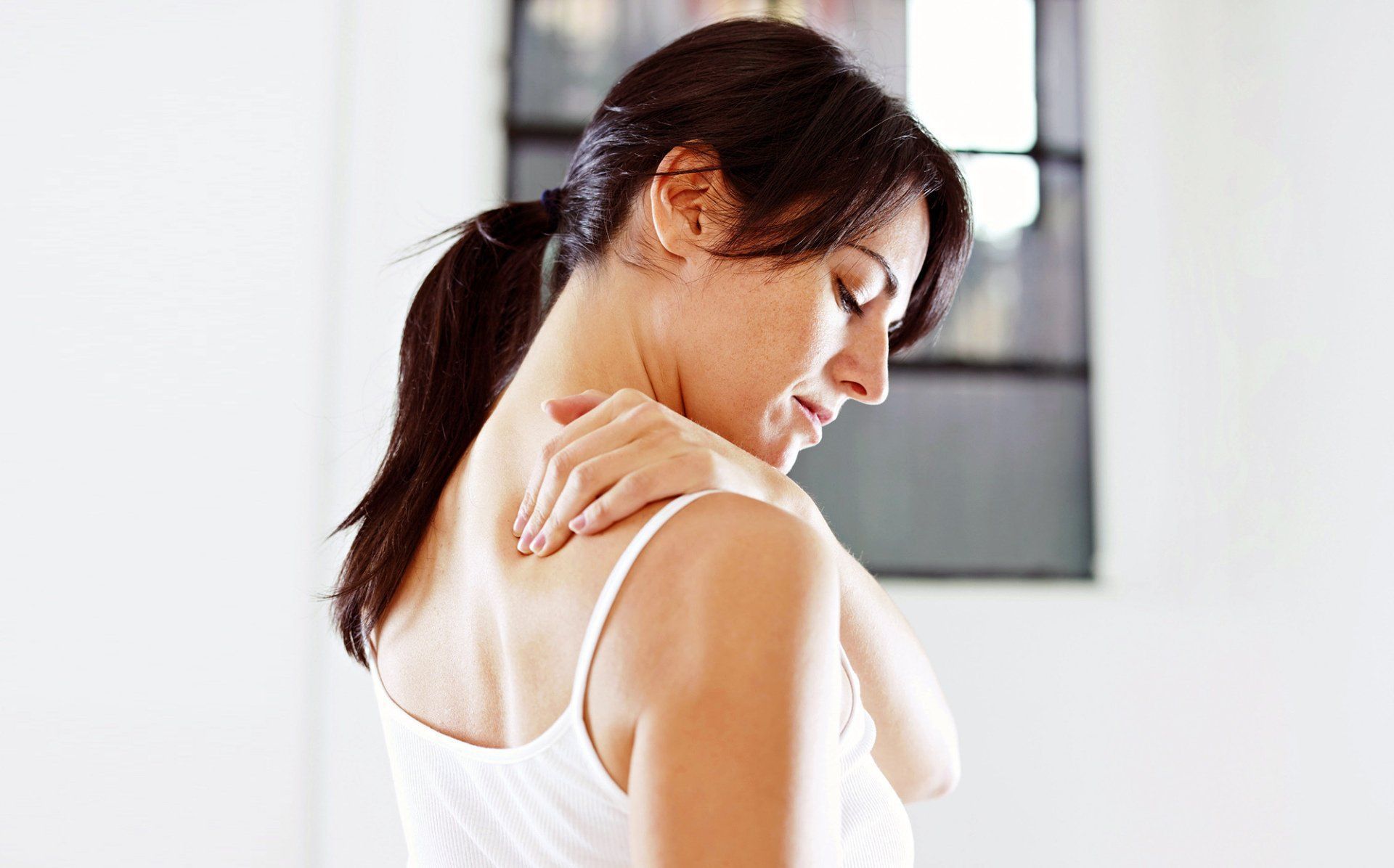 Woman in white top rubbing her shoulder, looking down in a bright indoor room.