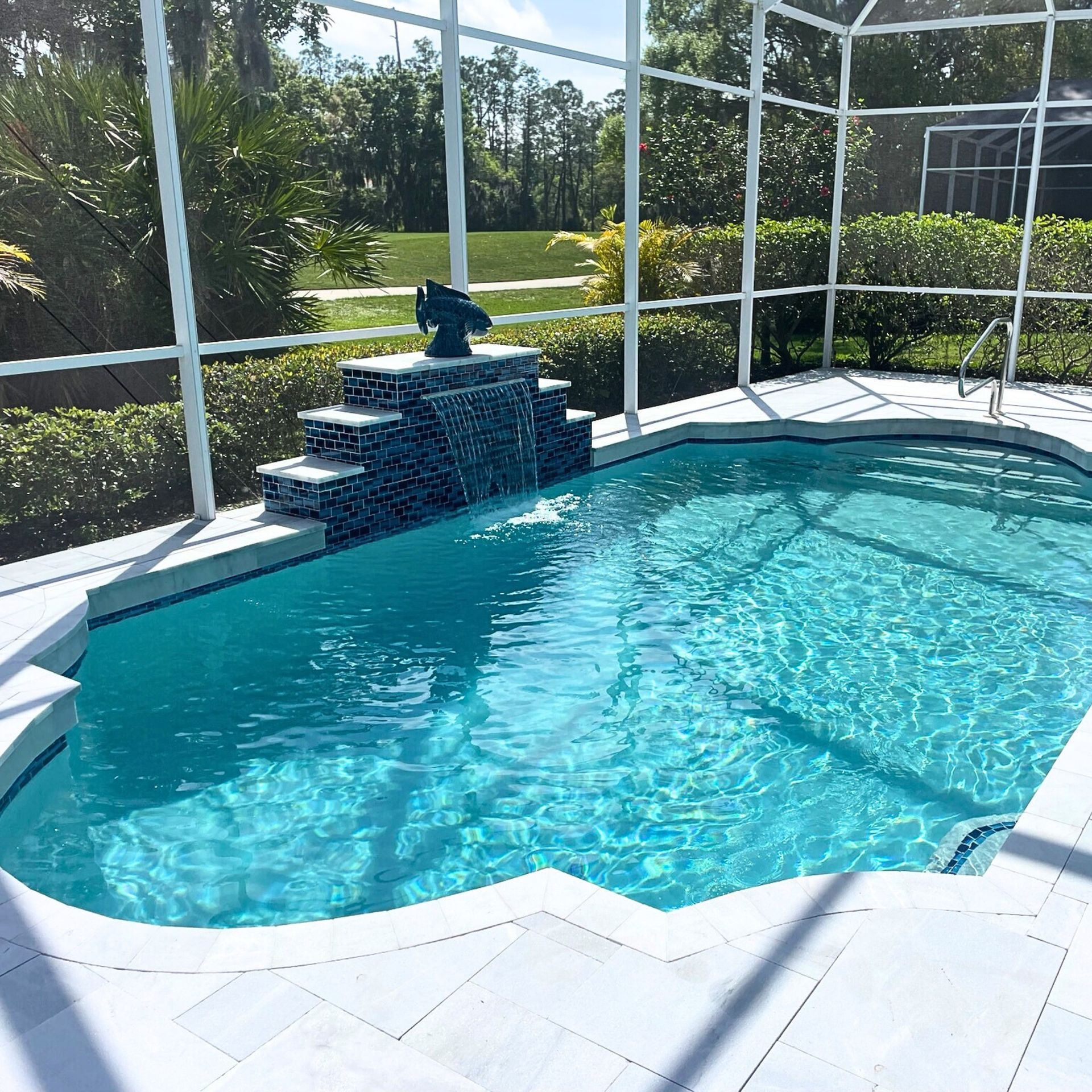 Swimming pool with waterfall feature, surrounded by screening, and greenery. Bright blue water and sunny day.