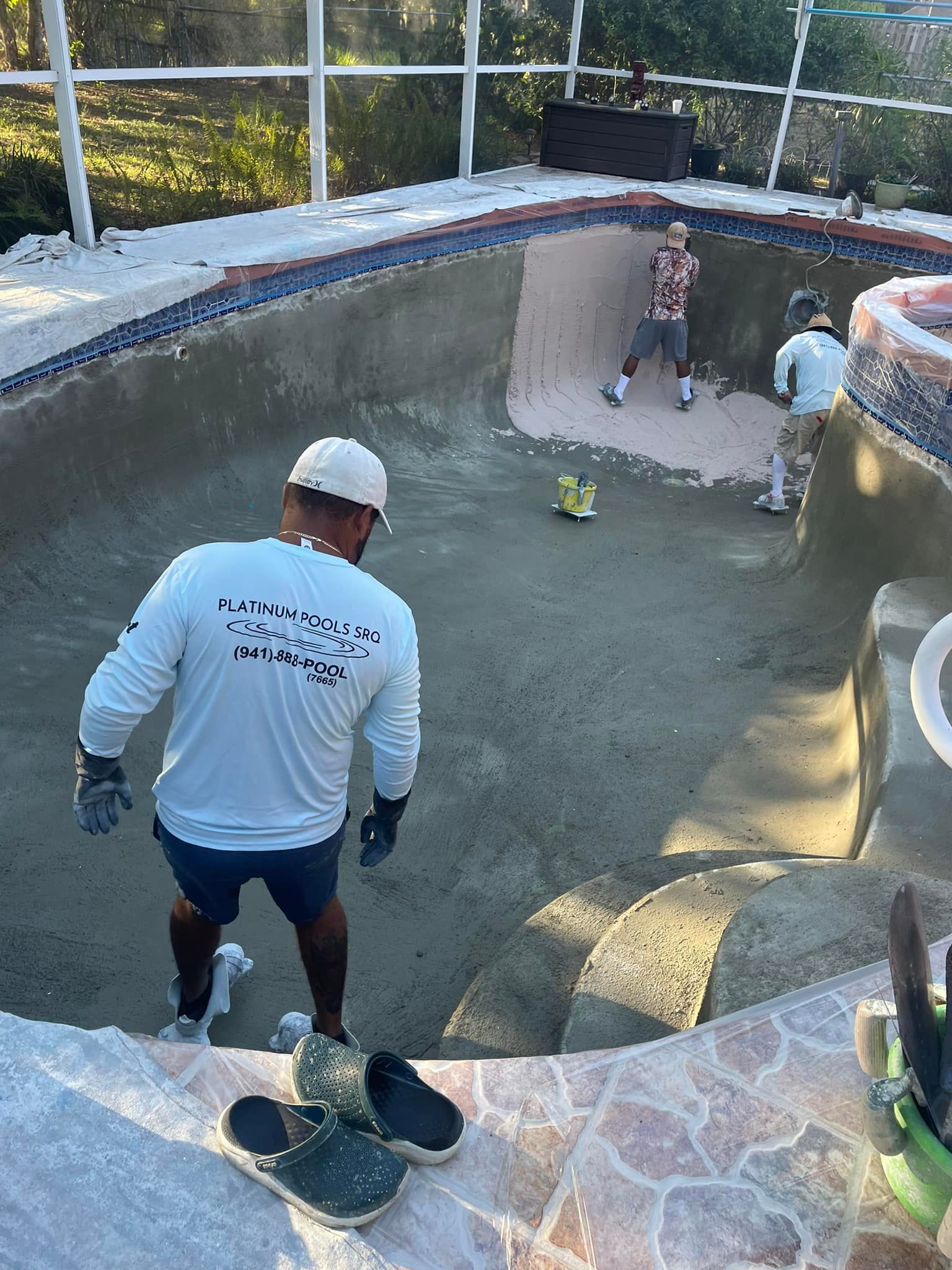 Men resurfacing a swimming pool. One enters the pool with tools and two others work from the edge.
