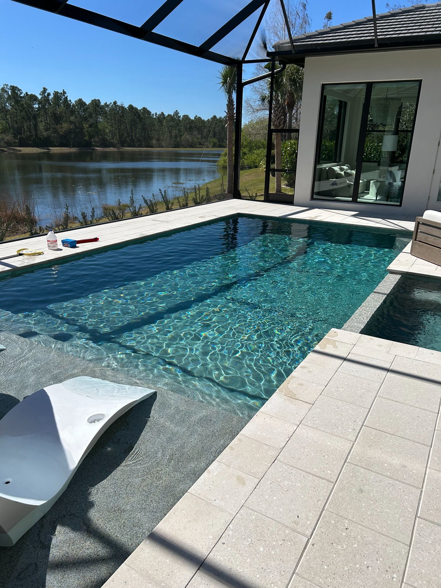 Poolside view with clear water, patio, lounge chair, and adjacent body of water.