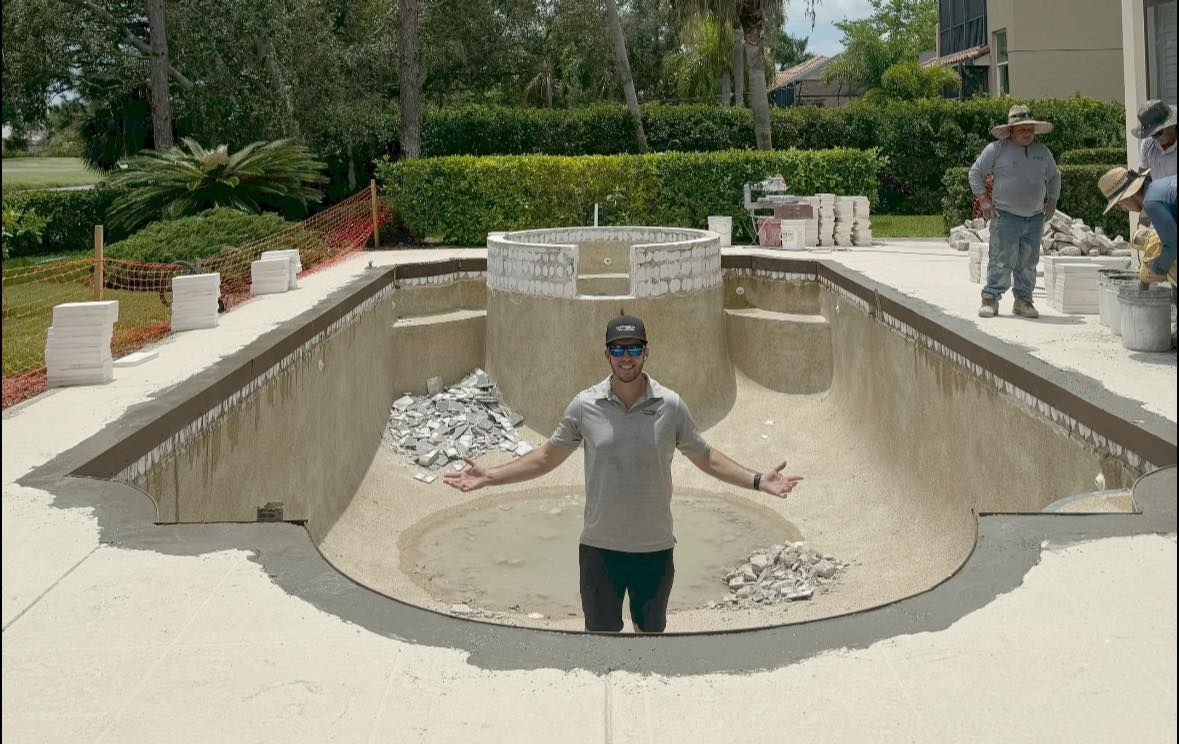 Man in a gray shirt stands in an empty pool, arms outstretched. Construction workers, trees, and a house are visible.