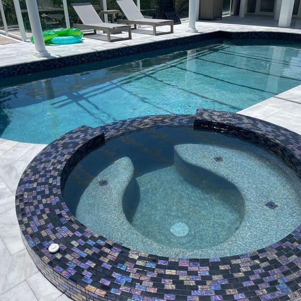 A pool and spa with blue mosaic tile, with lounge chairs in the background.