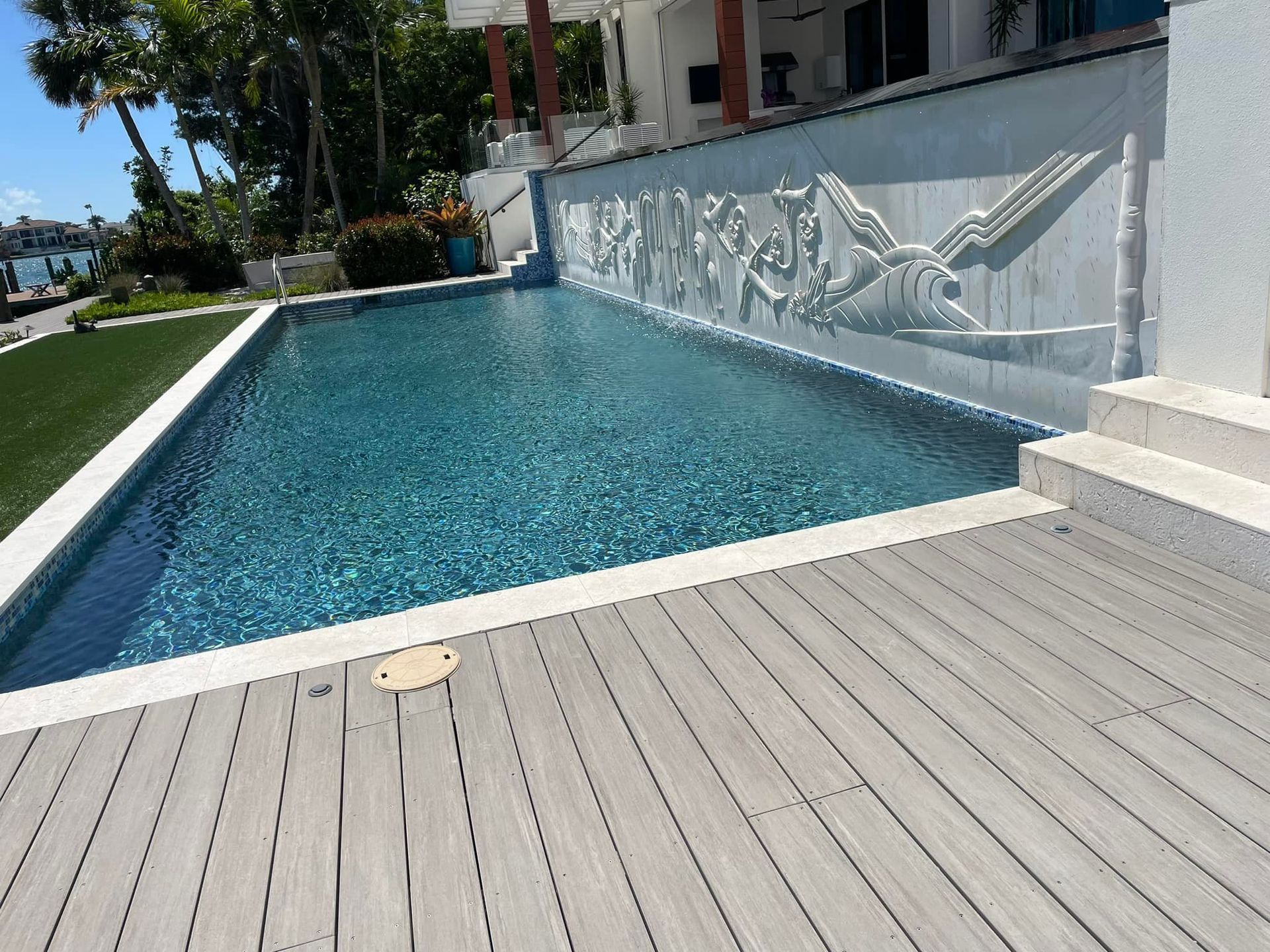 A rectangular swimming pool with dark blue water and a decorative wall feature, bordered by gray wooden decking.