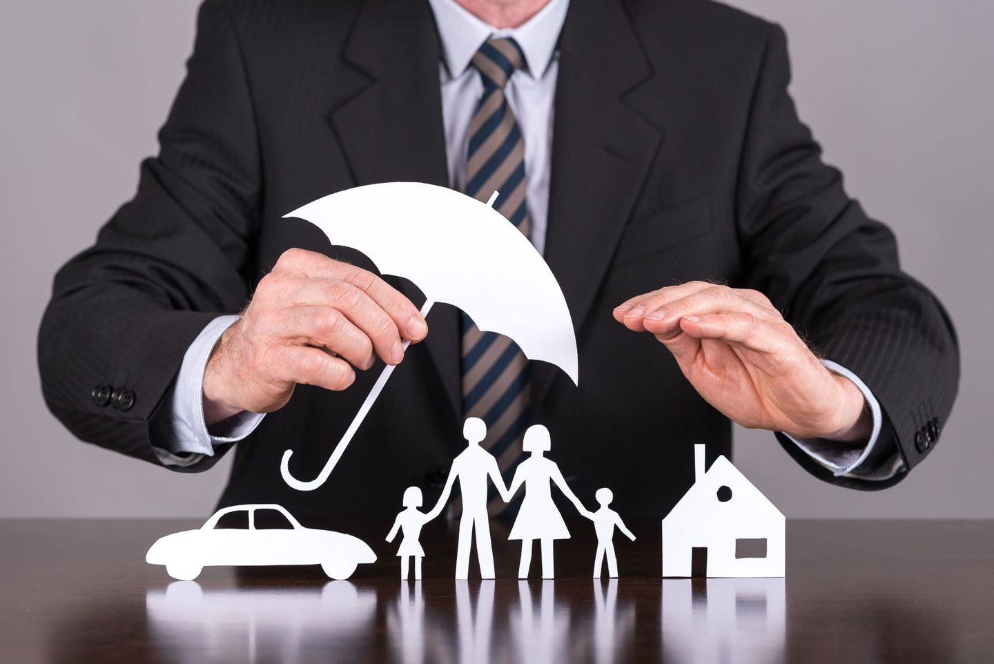 Businessperson in a suit holding protective umbrella cutout over paper figures representing a car, family, and house, symbolizing insurance coverage.
