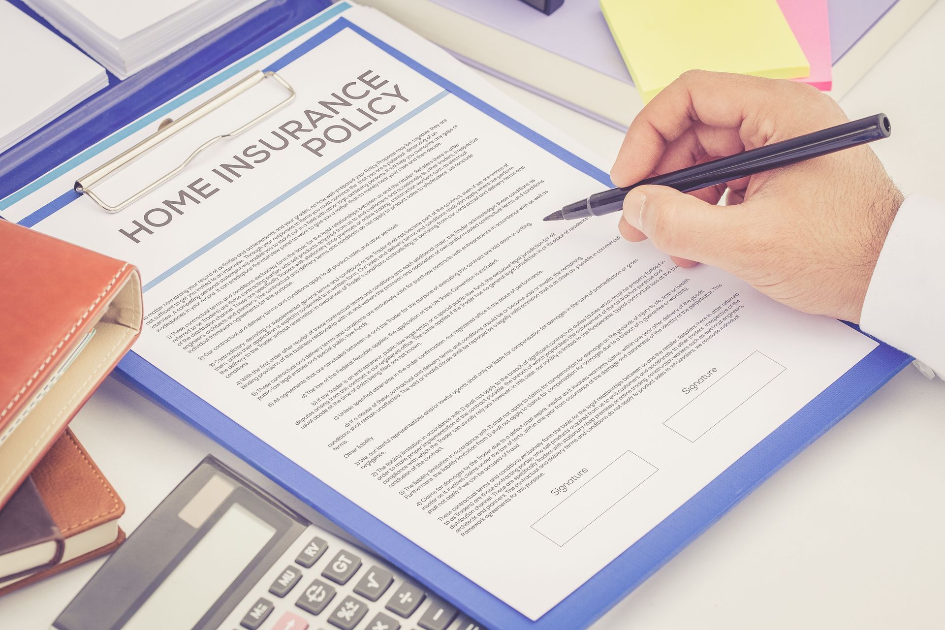 Person reviewing and preparing to sign a home insurance policy document on a clipboard, with a calculator and notebooks nearby.