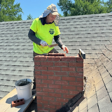 Worker repairing a red brick chimney on a shingled roof with a trowel and bucket nearby