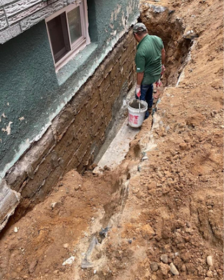 Worker inspecting a foundation trench beside a house wall with exposed concrete and dirt