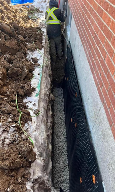 Worker in hi-vis vest standing in a narrow trench beside a brick wall, with excavation soil piled on the left.