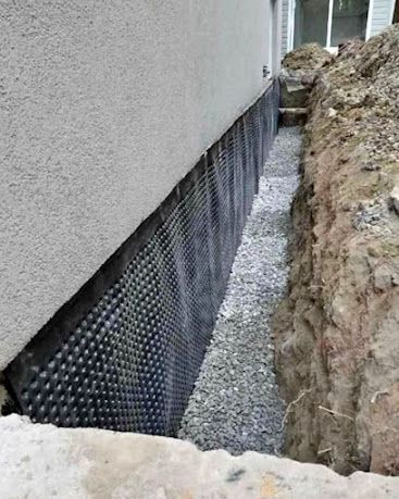 Foundation wall with black waterproofing membrane beside a narrow gravel trench and soil excavation