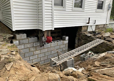 House lifted on cinder blocks over a dug-out foundation, with a ladder spanning the excavation.