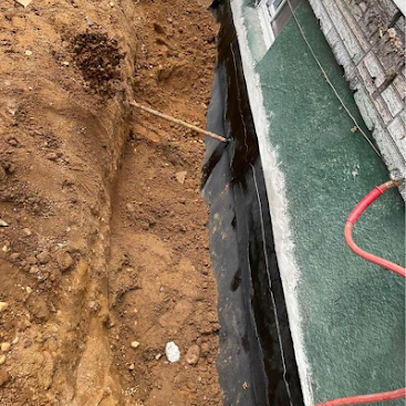 Trench beside a building foundation with exposed black waterproofing and dirt excavation