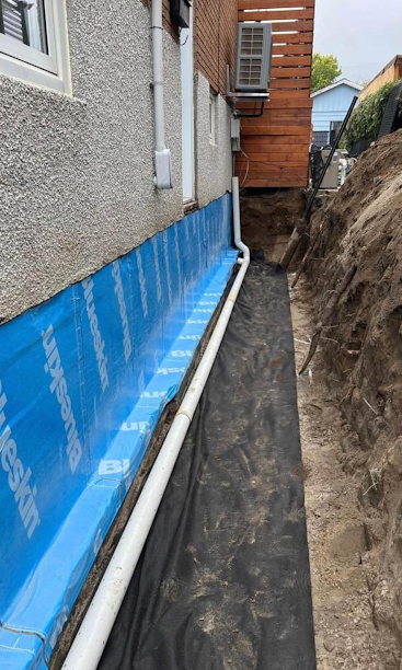 Narrow side yard with blue house wrap, exposed pipes, and a trench beside a building.