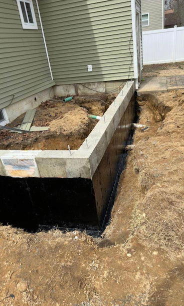 Concrete foundation wall beside a house, with exposed dirt and a black waterproofed lower section