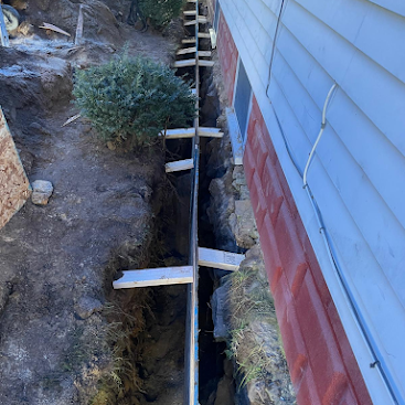 Narrow trench beside a house wall with a long board and wooden supports spanning it.