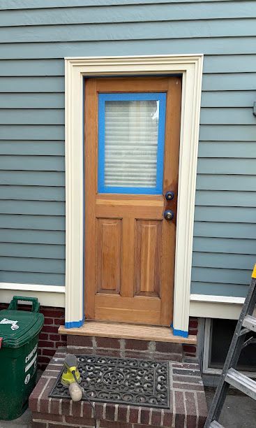 Front door with wood paneling and blue trim on a gray-sided house, with a doormat and bucket on the porch.