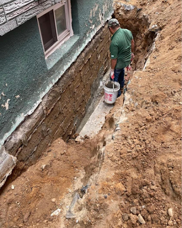 Worker in a trench beside a house, applying sealant to a foundation wall.