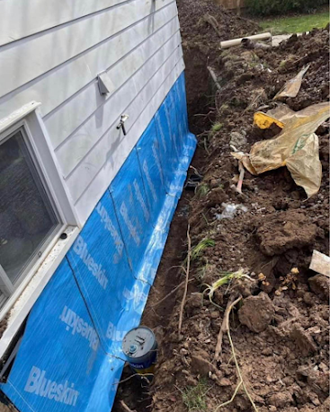 House wall with BlueSkin waterproofing, exposed dirt trench, and excavation debris along the foundation
