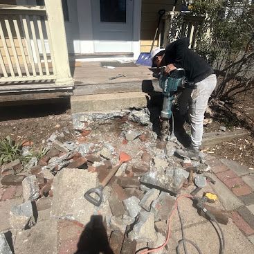 Worker using a jackhammer to break up concrete rubble beside a house porch