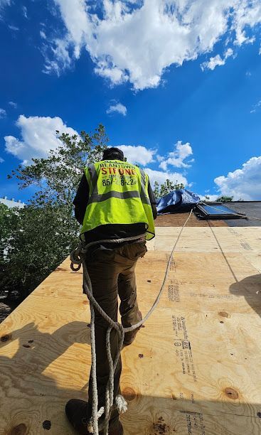 Worker in a neon vest climbs a roof with a safety harness under a blue sky.