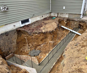 Foundation excavation beside a house, with rebar and concrete footings visible in a large dirt trench.