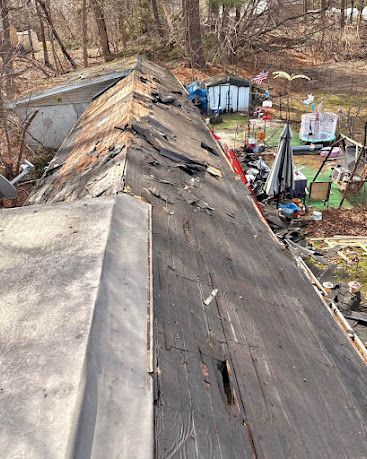 Damaged roof with peeling black shingles beside a cluttered yard and wooded area