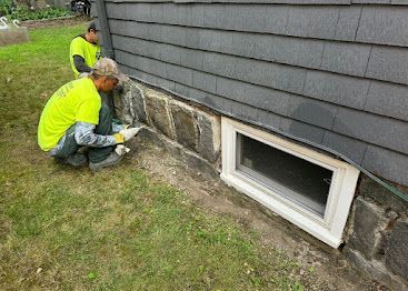 Workers in neon vests repair a home's stone foundation near a basement window.