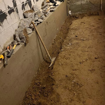 Narrow dirt trench beside a concrete wall, with rubble and a shovel leaning against it.