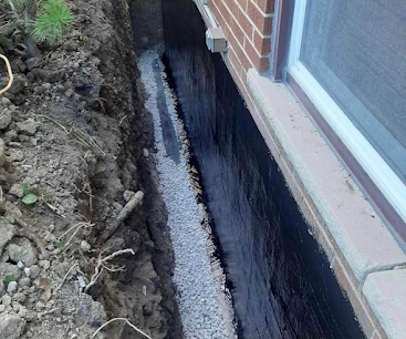 House foundation trench beside a brick wall, showing exposed black waterproofing and gravel
