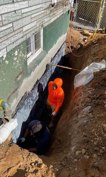 Two workers in a deep trench beside a brick house, one in an orange hoodie near exposed foundation.