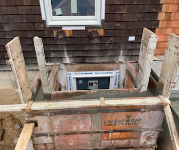 Basement window well beneath a shingled house, framed by wooden supports and construction debris.