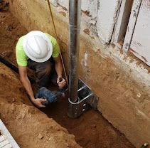 Worker in white hard hat installing a metal post in a dirt trench beside a wall.