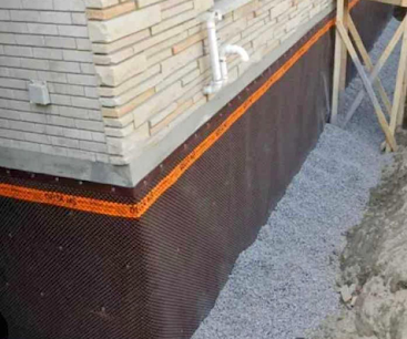 Foundation wall with black waterproofing membrane and orange drainage strip beside gravel ground