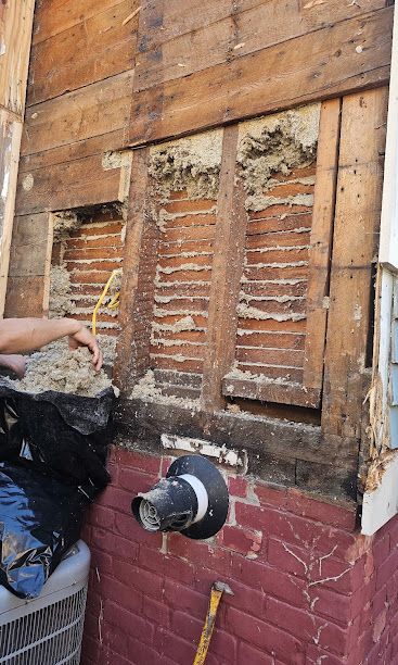 Water-damaged wall with broken wooden shutters and a protruding pipe, while someone clears debris nearby