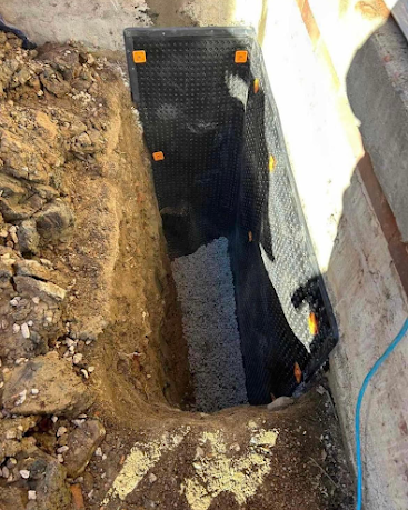 Narrow excavation beside a wall, lined with black waterproof membrane with orange fasteners and muddy soil around it