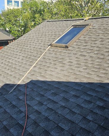 Rooftop with a solar panel and cable on gray shingles, partly shaded by a nearby tree