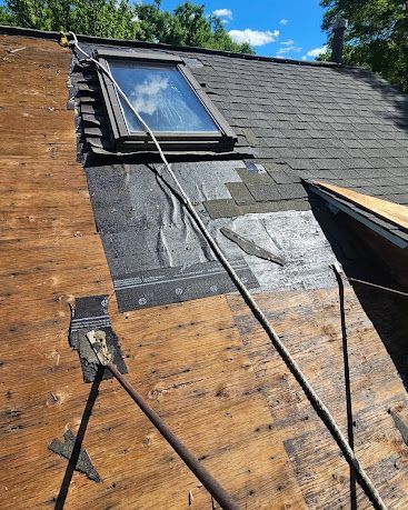 Roof repair with exposed plywood, shingles, and a skylight under a clear blue sky