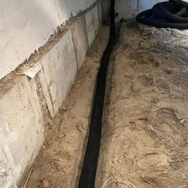 Black drainage pipe laid in a sandy trench beside a concrete wall.
