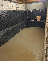 Empty indoor stall with black walls and a muddy floor, lit by overhead lights.