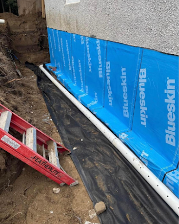 Blue exterior wall wrap over black sheeting beside a ladder and white pipe in a narrow dirt trench