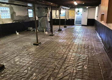 Large unfinished basement with exposed beams, support posts, and a tiled floor, lit by small windows and ceiling lights