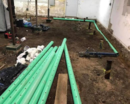 Trenched construction site with green PVC pipes laid over muddy ground beside a building wall
